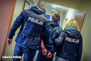 Policjanci prowadzą zatrzymanego z założonymi kajdankami na ręce trzymane z tyłu