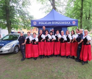 Pamiątkowe zdjęcie zespołu wokalno- tanecznego wraz z policjantami przy profilaktycznym stoisku policyjnym.