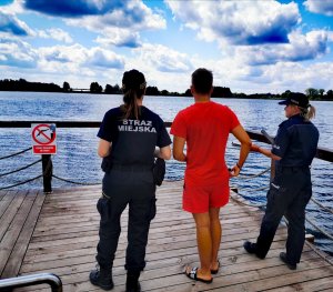 Na zdjęciu policjantka, funkcjonariuszka Straży Miejskiej oraz Ratownik stoją na pomoście przy plaży.